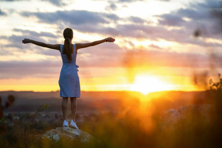 A young woman in summer dress standing outdoors with outstretched arms enjoying view of bright